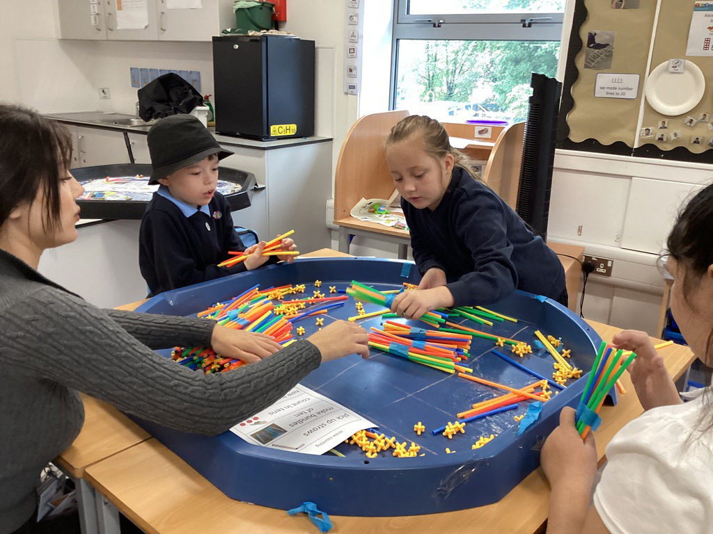 Four children and an adult sit around a blue activity table, playing with colourful plastic straws and connectors. The classroom setting is bright, with large windows and educational materials on the walls.