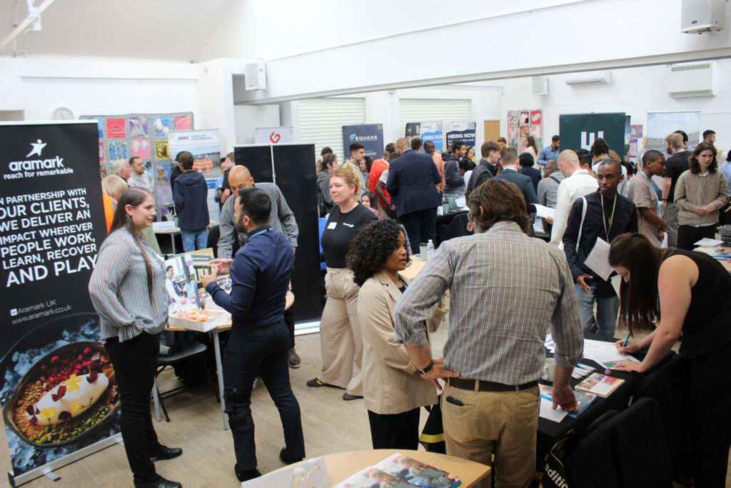 A busy careers fair with people networking at various company stands, engaging in conversations, handing out leaflets, and exchanging information in a bright, indoor setting.