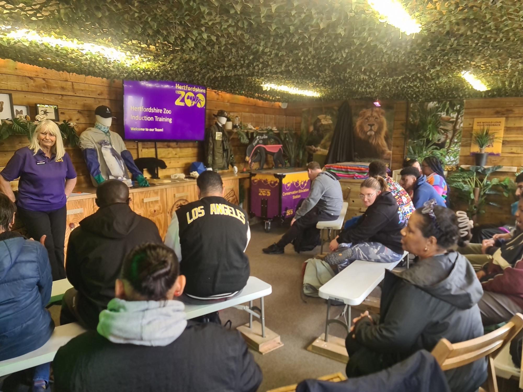 A group of people sits on benches indoors, facing a screen displaying “Hertfordshire Zoo Education Training.” A woman in a purple shirt stands in front, and the room is decorated with wooden walls, plants, and animal figures.