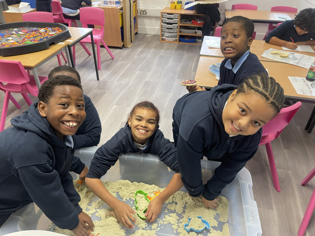 Four children smile and play with sand and toy moulds in a classroom. Pink chairs, tables, and shelves with art supplies are visible in the background.