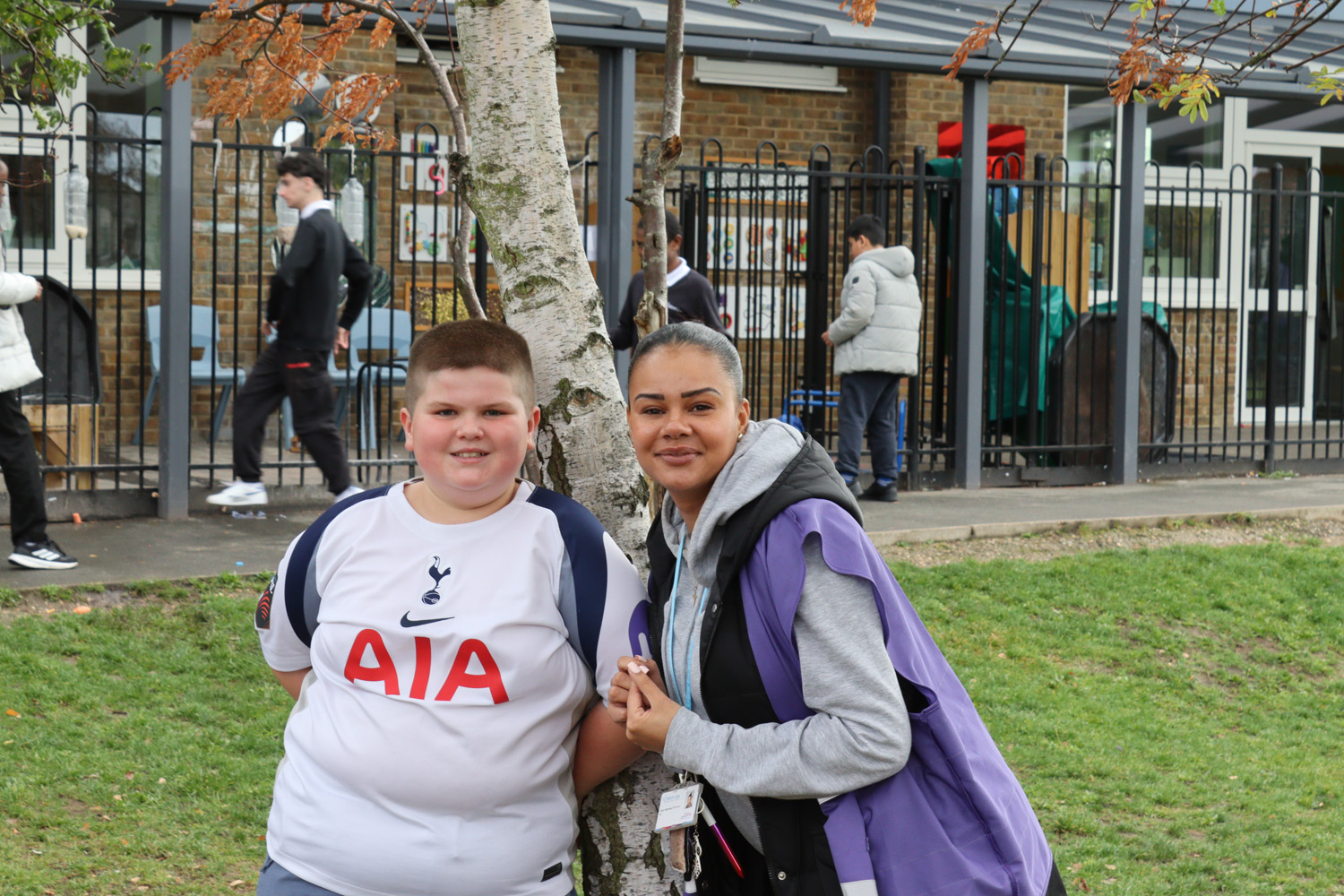 A boy in a white Tottenham Hotspur shirt and a woman in a purple waistcoat smile together by a tree on a grassy area, with a school building and other pupils in the background.