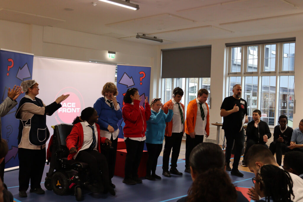 A group of people, including one in a wheelchair, stand on a stage indoors, some applauding. Others act or speak. An audience watches. Colourful costumes, props, and a large screen with “RACE FRONT” are visible in the background.