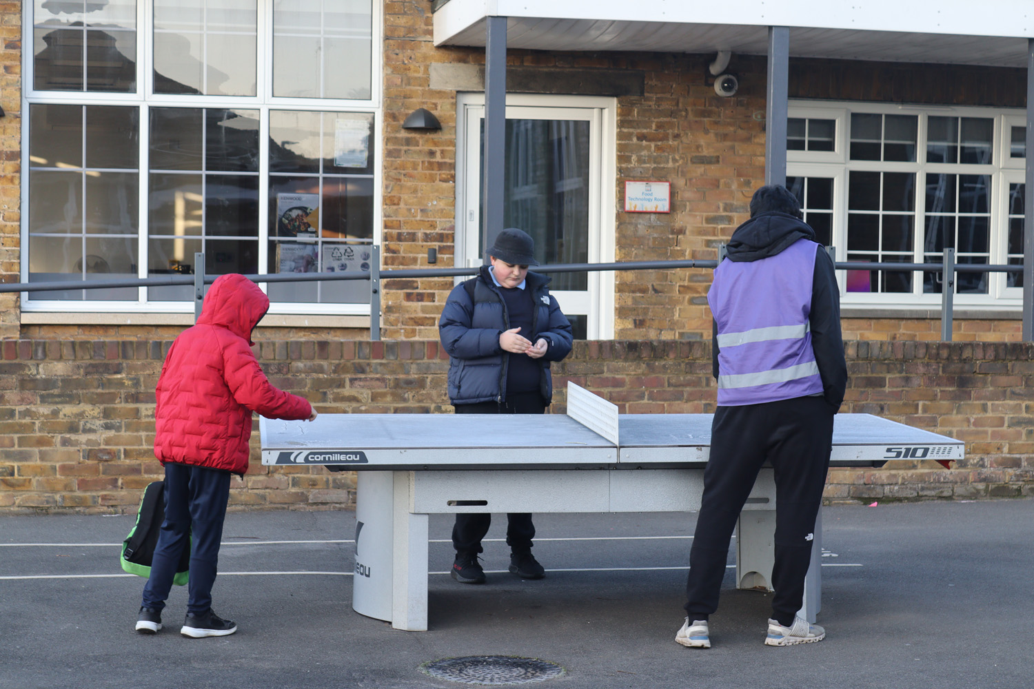 Three people are near an outdoor table tennis table. One child in a red jacket holds a bat, while an adult in a purple waistcoat stands on the other side. Another adult in a dark coat looks down at a mobile phone.