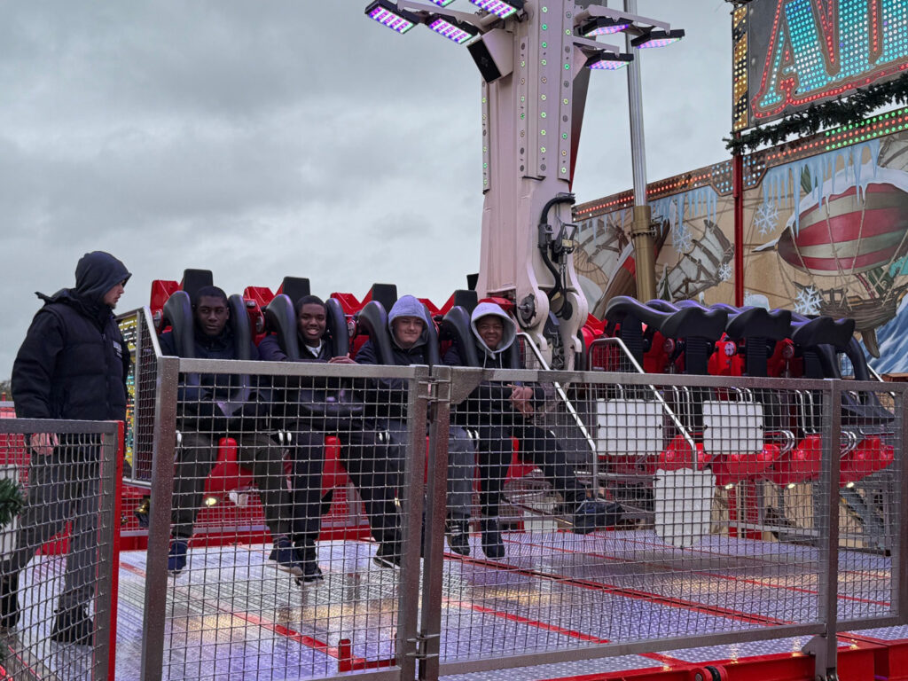 Five people sit in seats on an amusement park ride, secured with harnesses, smiling and posing for a photo. A person in a hooded jacket stands nearby. The background shows colourful lights and a mural.
