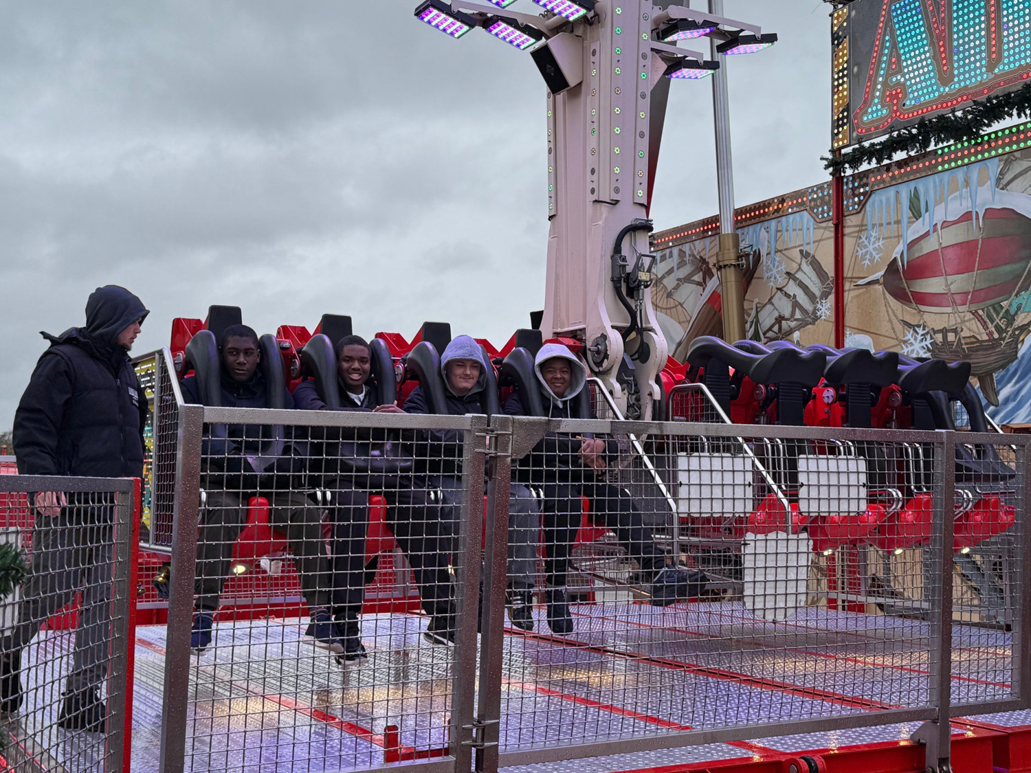 Five people sit in seats on an amusement park ride, secured with harnesses, smiling and posing for a photo. A person in a hooded jacket stands nearby. The background shows colourful lights and a mural.