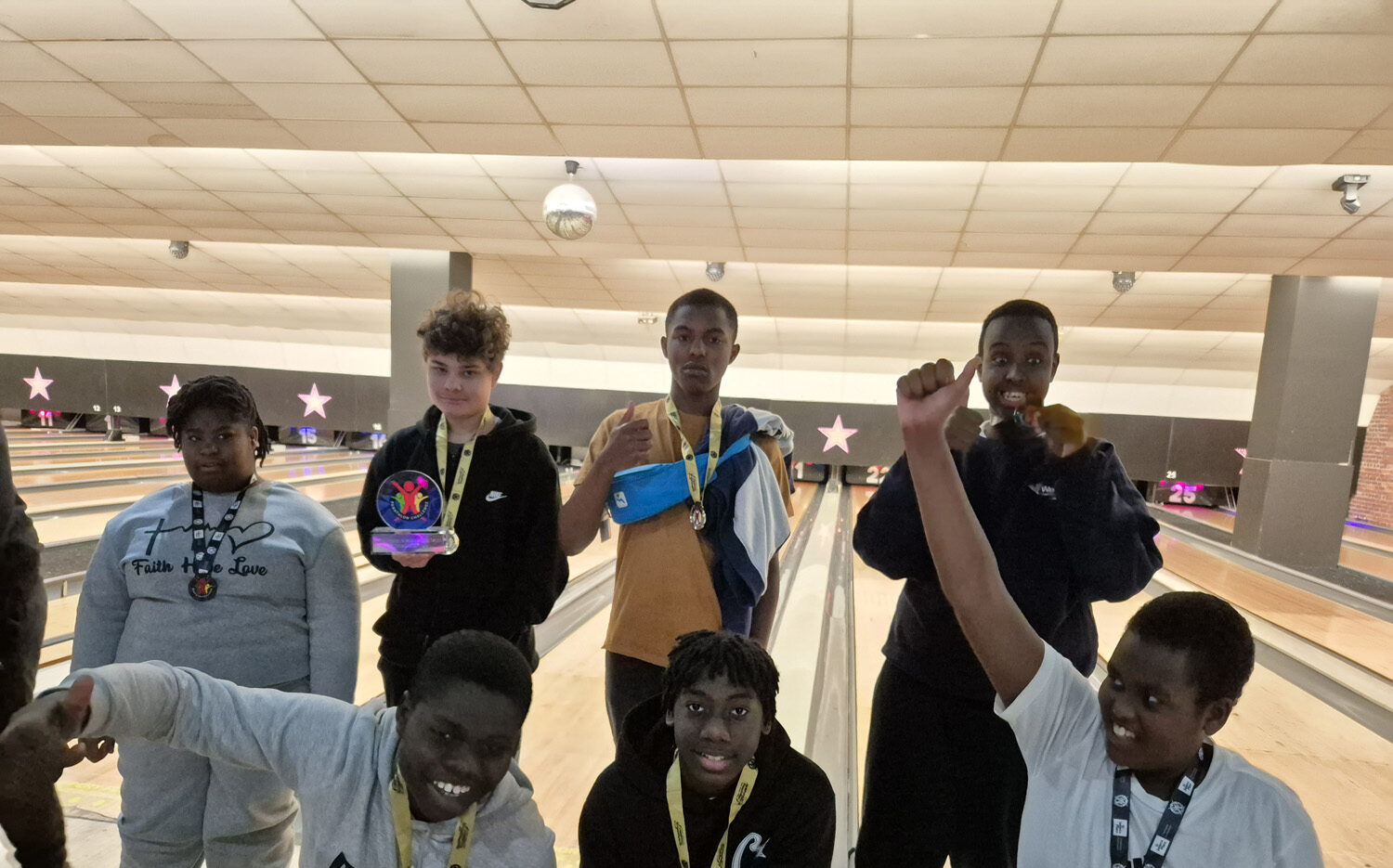 Seven boys pose and smile in front of bowling lanes, some wearing medals round their necks. One boy holds a trophy, and another raises his fist in celebration. The background shows pins and lanes under bright lights.