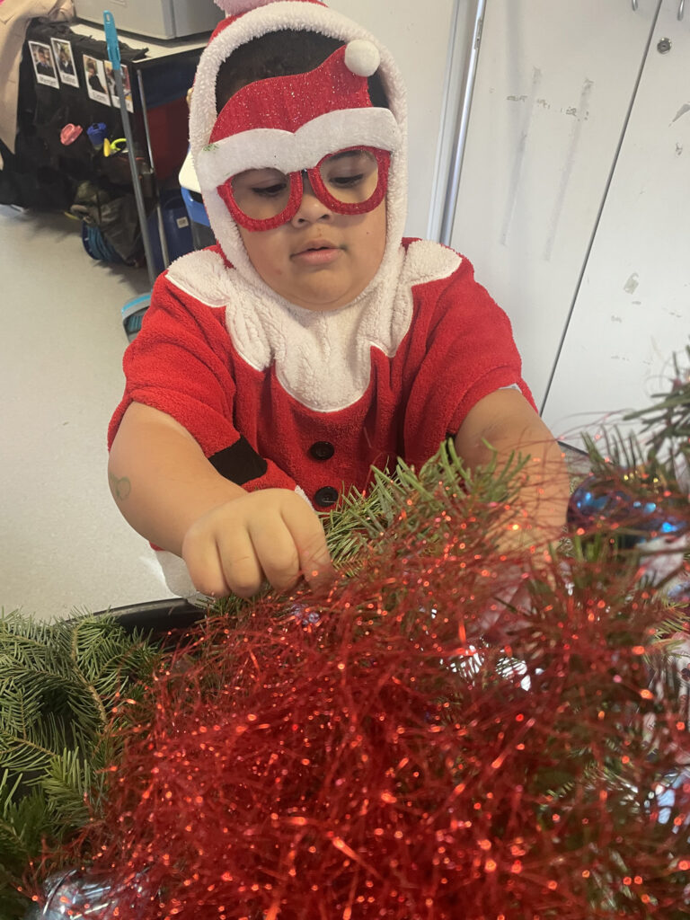 A child from West Lea School, dressed in a Father Christmas costume and red spectacles, decorates a Christmas tree with red tinsel and green branches indoors.