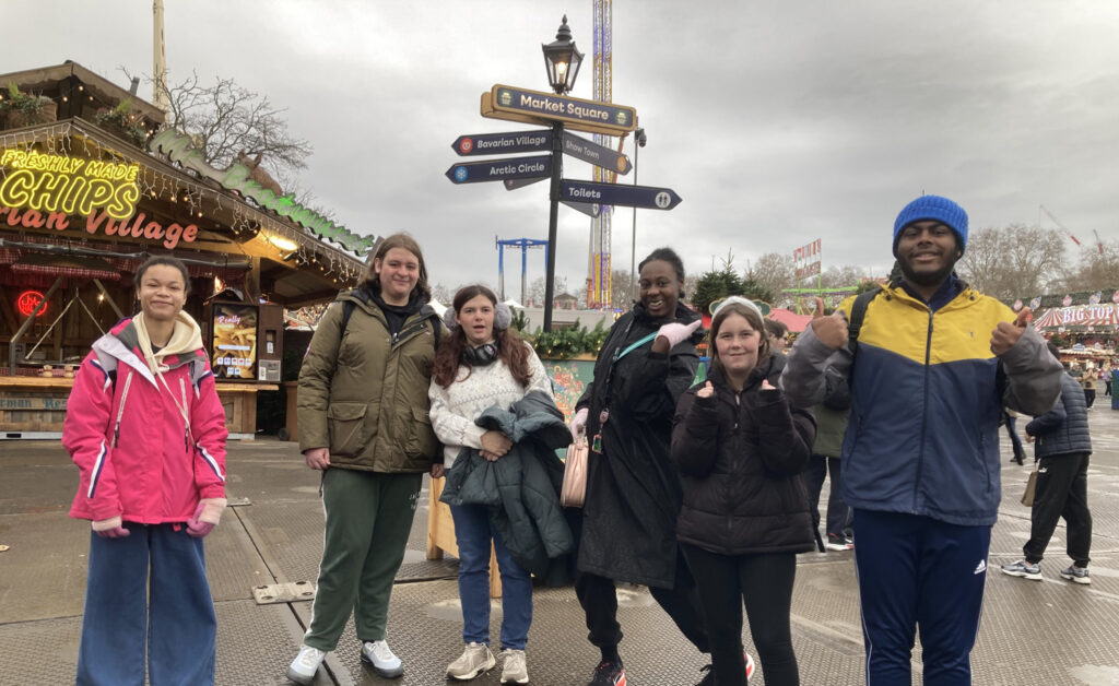 Six people stand and pose together at an outdoor fair or winter market, with festive lights, a food stall, and several signposts in the background under a cloudy sky.