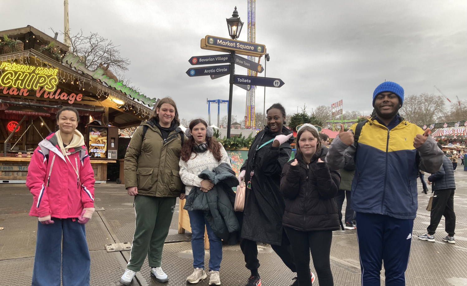 Six people stand and pose together at an outdoor fair or winter market, with festive lights, a food stall, and several signposts in the background under a cloudy sky.