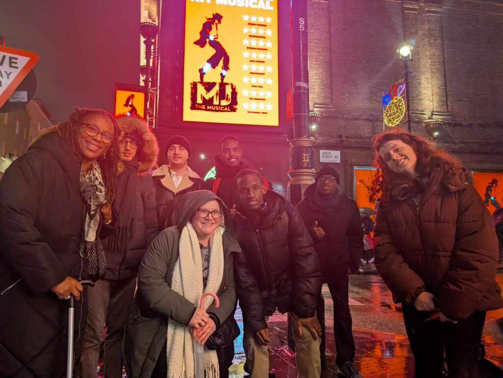 A group of eight people in winter clothing pose and smile on a wet city street at night with bright neon signs behind them, including a large sign for MJ The Musical.