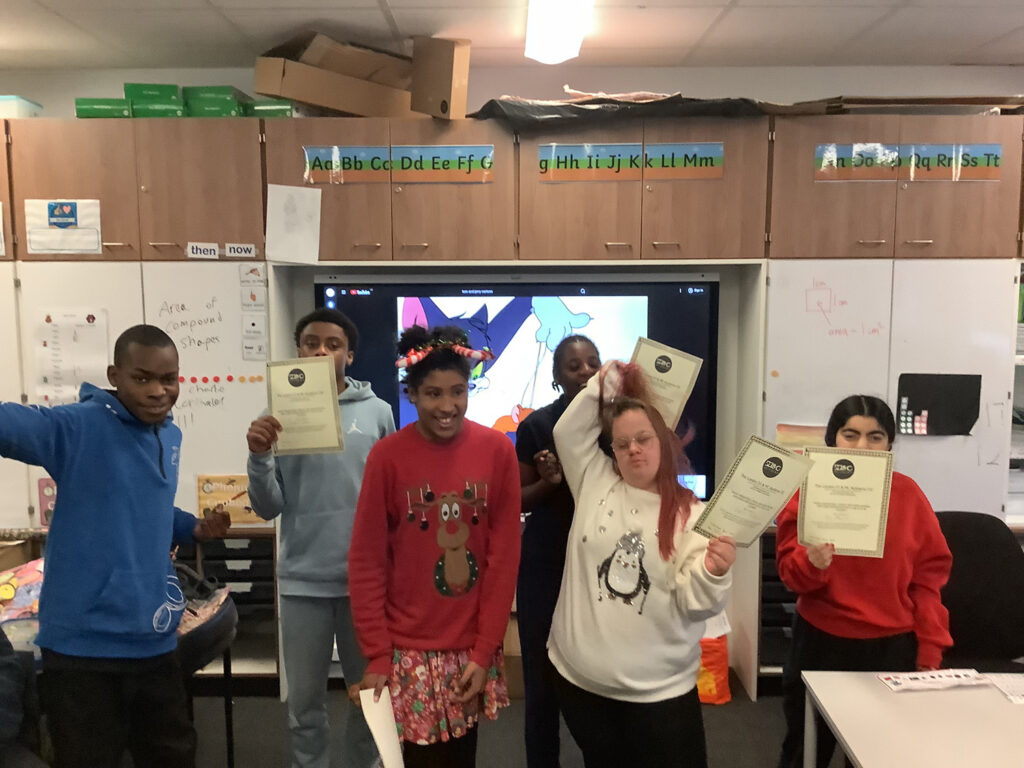 Six students stand in a classroom, smiling and holding certificates. They wear colourful jumpers, and a cartoon cat is displayed on the screen behind them. The classroom is decorated with alphabet labels and storage cupboards.