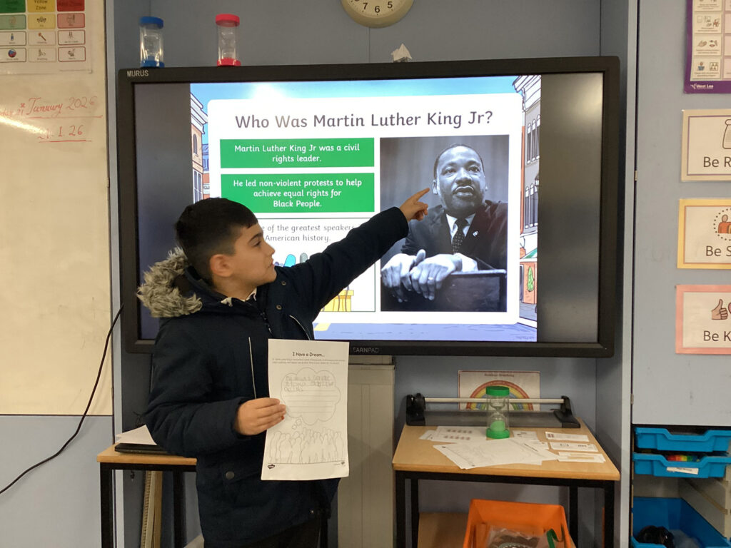 A young boy stands in front of a screen displaying information about Martin Luther King Jr., pointing to a black-and-white photo of King and holding a worksheet whilst presenting in a classroom.