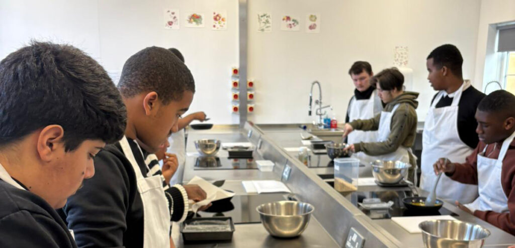 A group of students wearing aprons work at kitchen stations, reading recipes and preparing food in a bright, modern cookery classroom. Mixing bowls and cooking utensils are on the worktops.