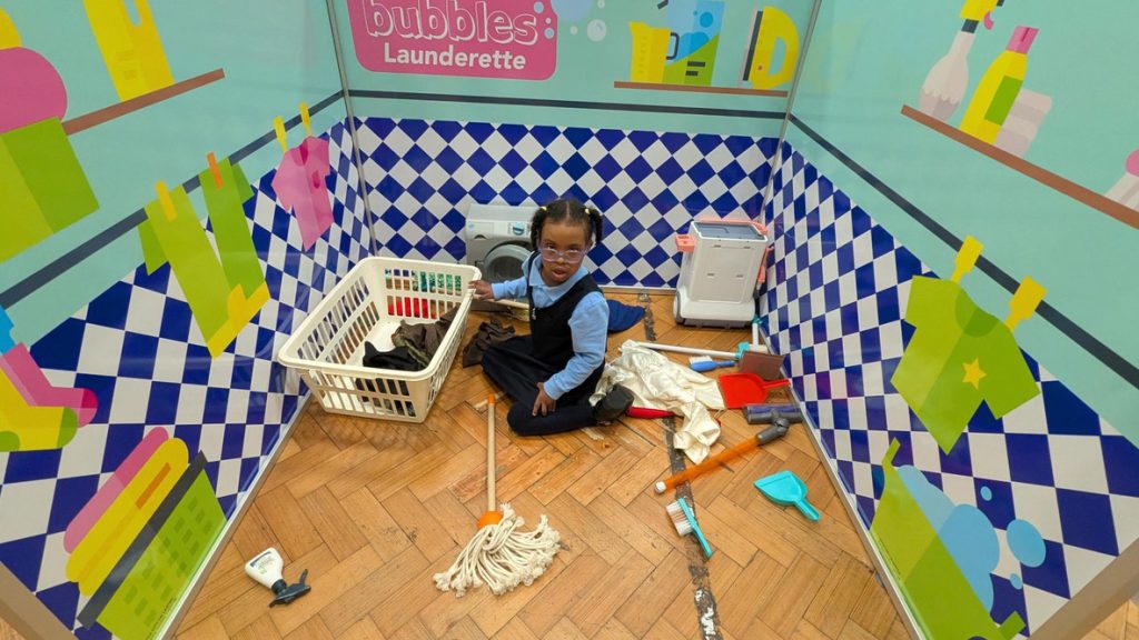 A child sits on the floor in a play launderette area with toy cleaning items, a laundry basket, and mop, surrounded by colourful walls decorated with cleaning supply illustrations.