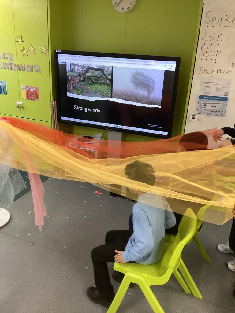 Two children sit on green chairs in a classroom, holding up orange and yellow translucent fabric. Behind them, a screen displays images of trees in strong winds with the text Strong winds.