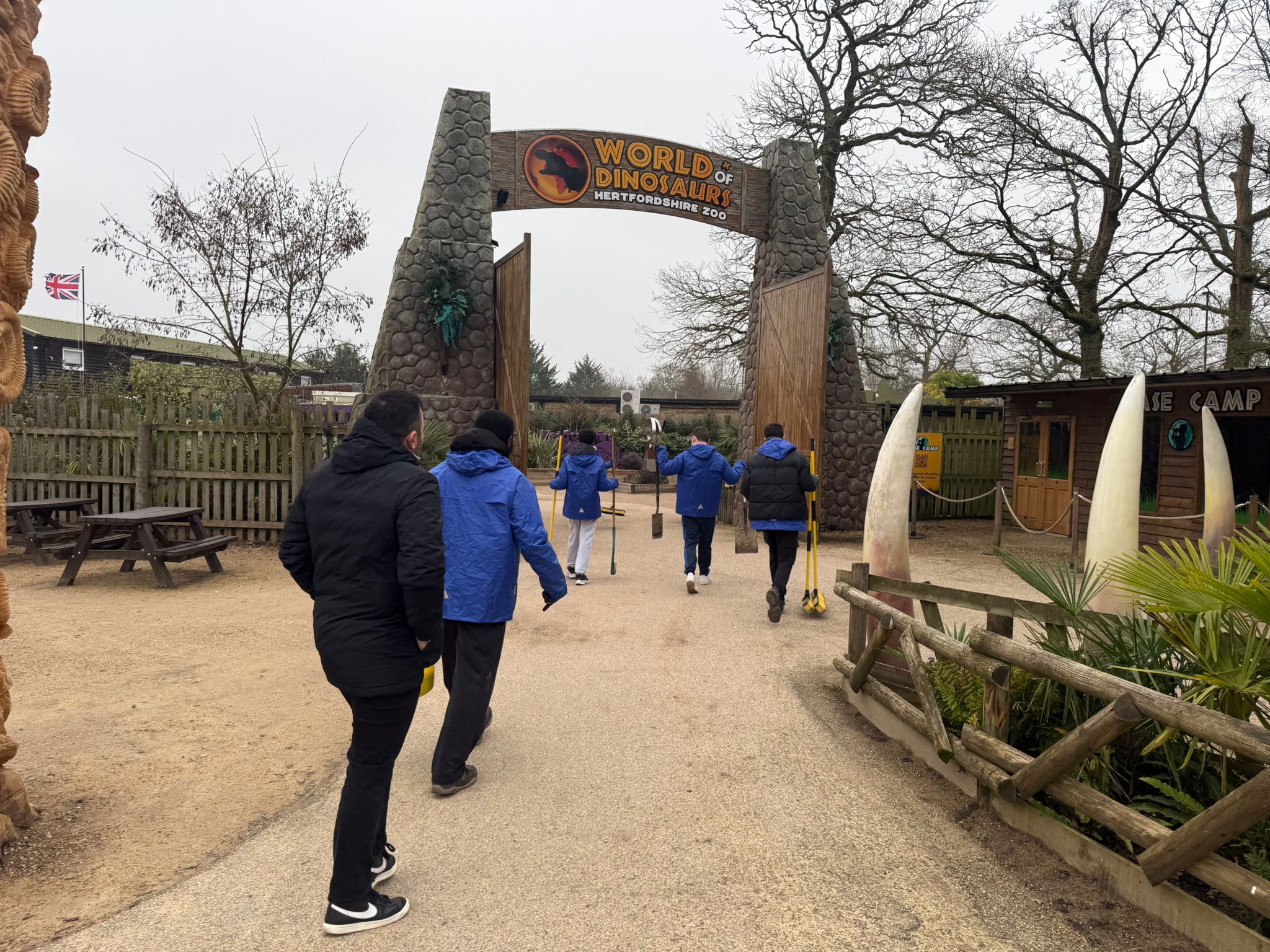 Several people wearing blue and black jackets walk towards the entrance gate of World of Dinosaurs at Hertfordshire Zoo on a cloudy day, surrounded by trees and wooden fences.