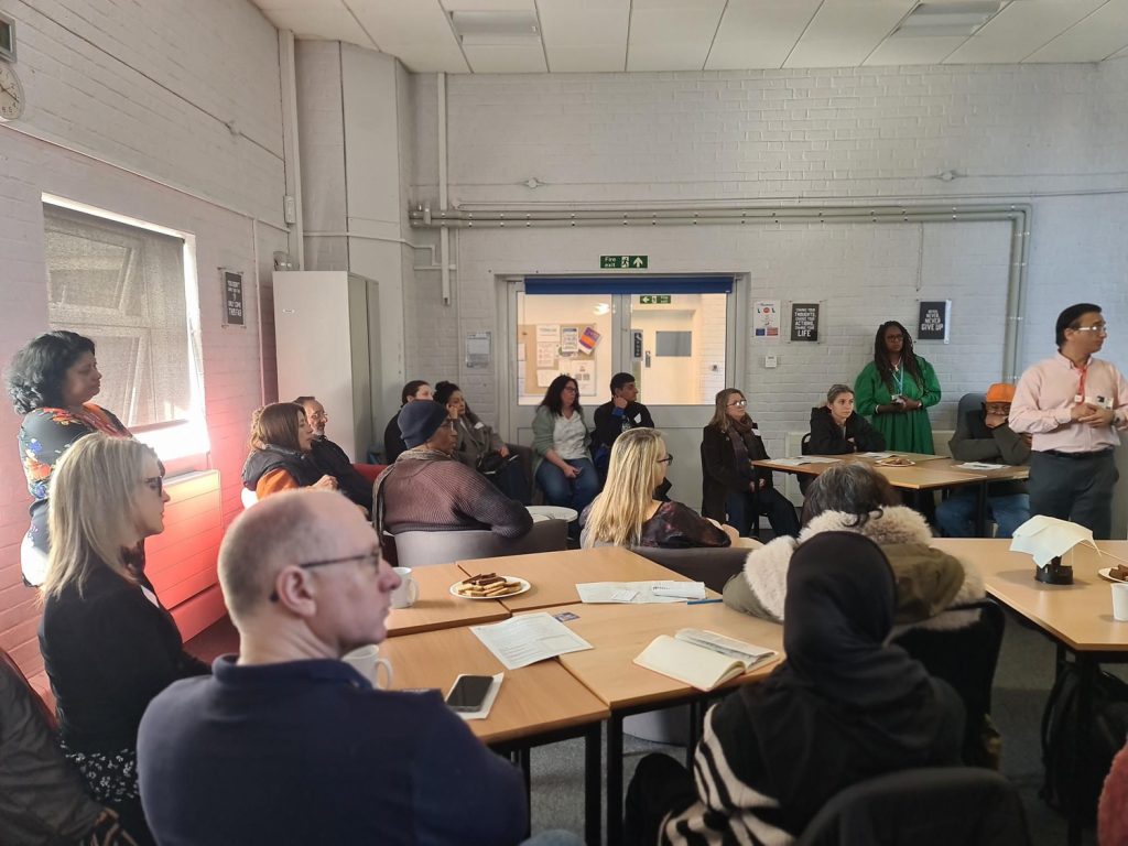 A group of people sit around tables in a brightly lit room, attentively listening to a person standing and speaking at the front. Some are taking notes, and snacks are on the tables.
