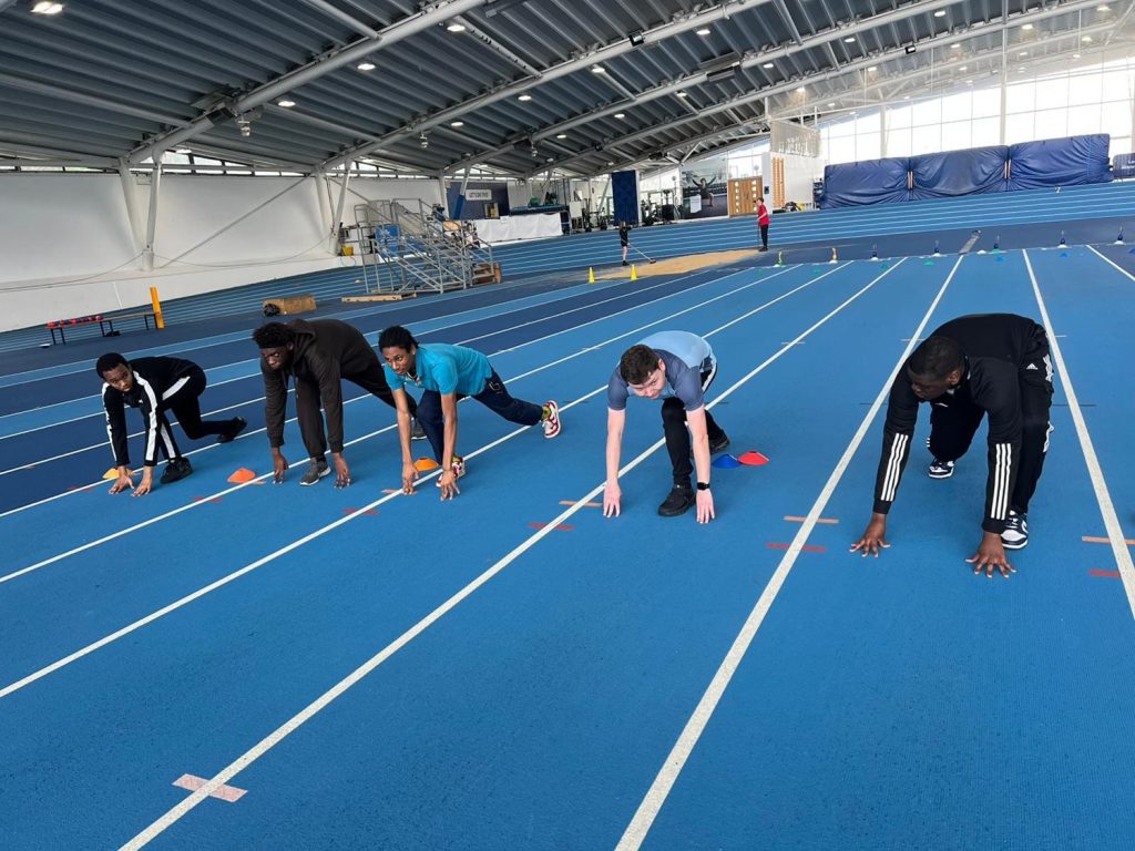 Five people are in starting positions on a blue indoor running track, preparing to sprint. The large indoor facility is bright, with seating and equipment visible in the background.