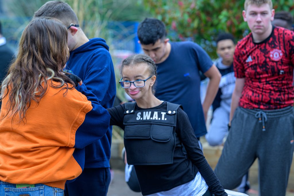 A young girl wearing glasses and a S.W.A.T. vest smiles and poses with her arm raised among a group of teenagers outdoors. Other children, dressed in casual clothes, stand and interact around her.