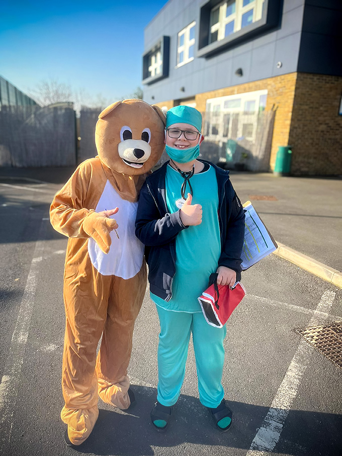 A person in a bear costume stands next to a person dressed as a doctor outside West Lea School. Both are smiling; the doctor holds documents and a red bag, giving a thumbs-up.