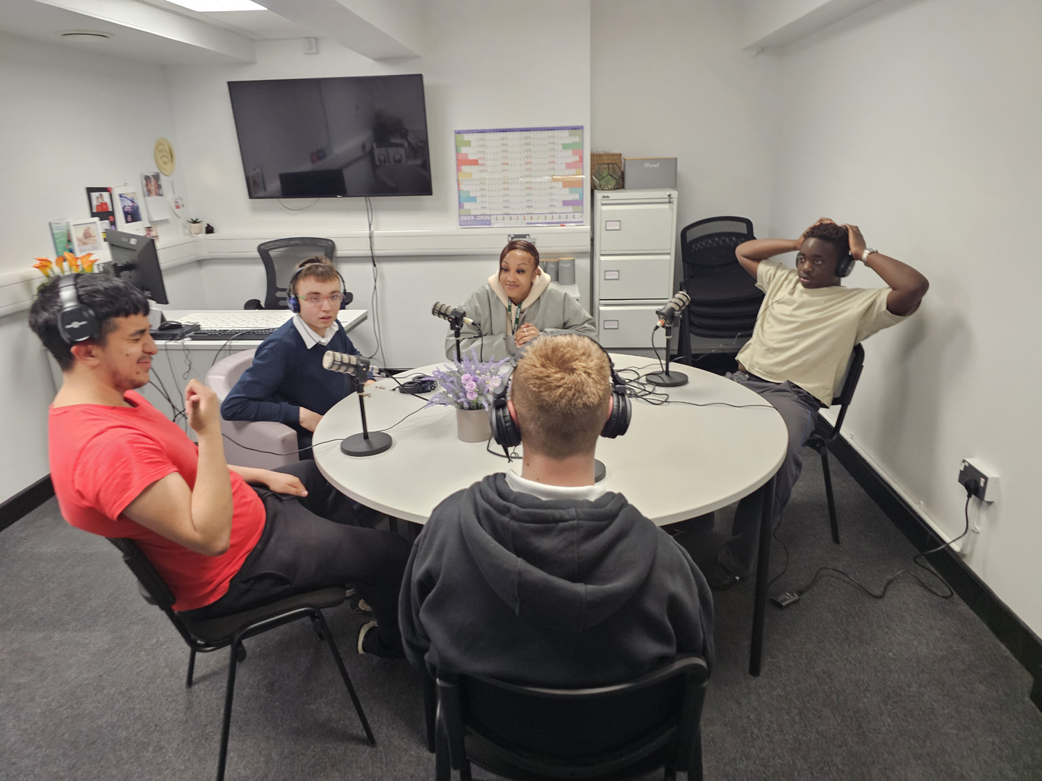 Five people are sitting around a round table with microphones, appearing to record a podcast in a modern office space with a TV, whiteboard, and filing cabinets in the background.