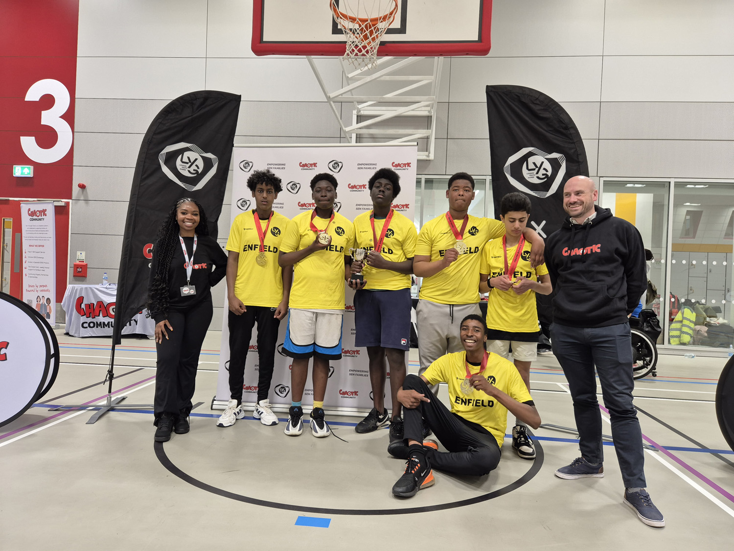 A basketball team of six boys in yellow jerseys pose with their trophies and medals, flanked by two adults. They stand in a gymnasium under a basketball hoop, with black flags and banners in the background.