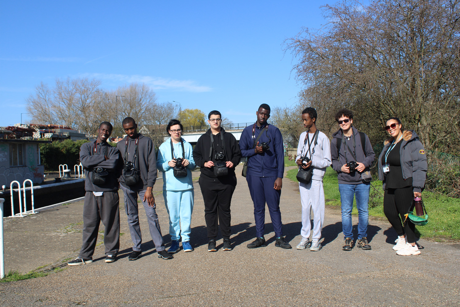 A group of eight people stand side by side outdoors on a sunny day, each holding a camera. Trees and a blue sky are in the background. Most are wearing casual clothes and sunglasses.