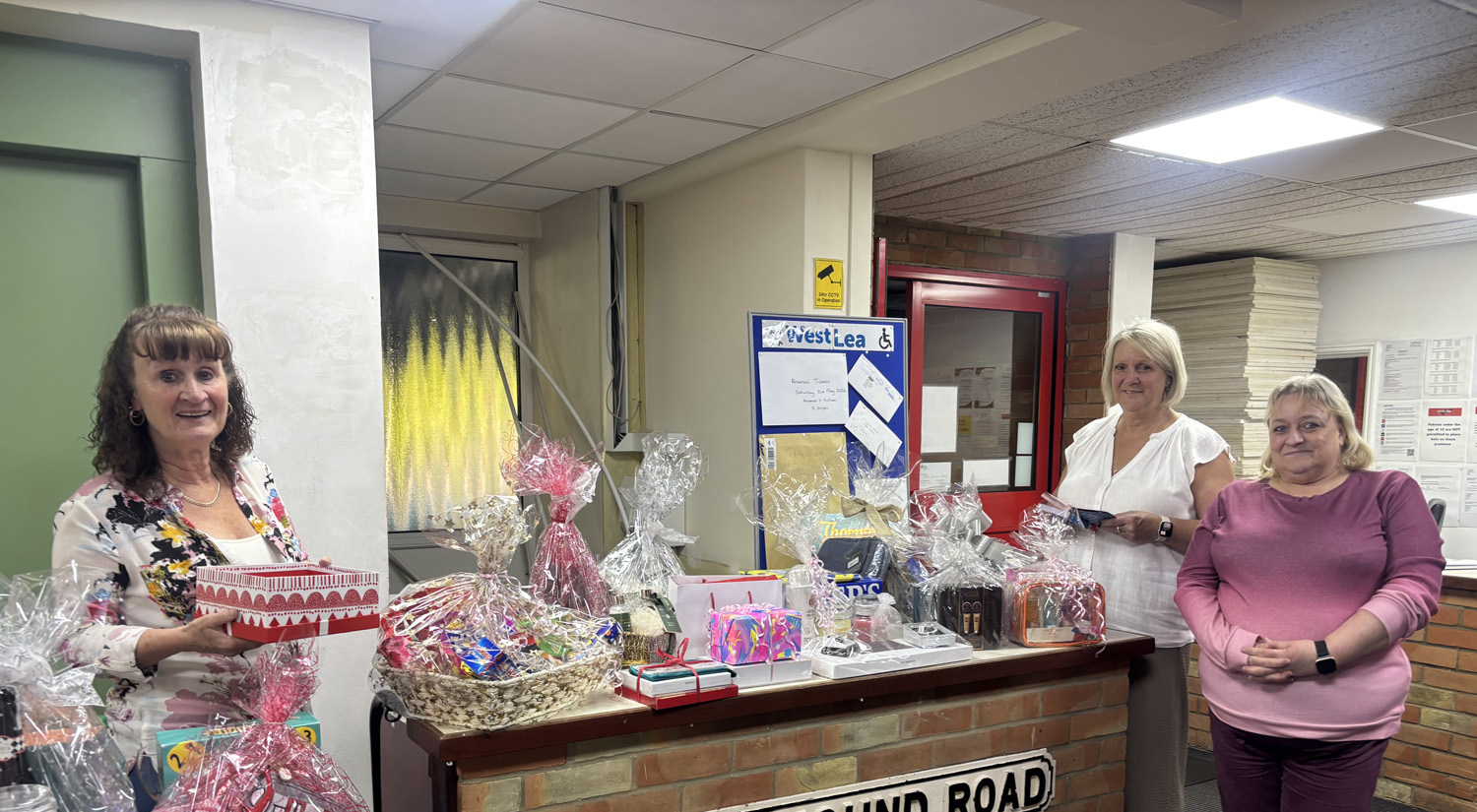 Three women stand behind a reception desk covered with wrapped gift hampers and boxes. They are smiling and appear to be in an office or community centre with noticeboards and papers in the background.