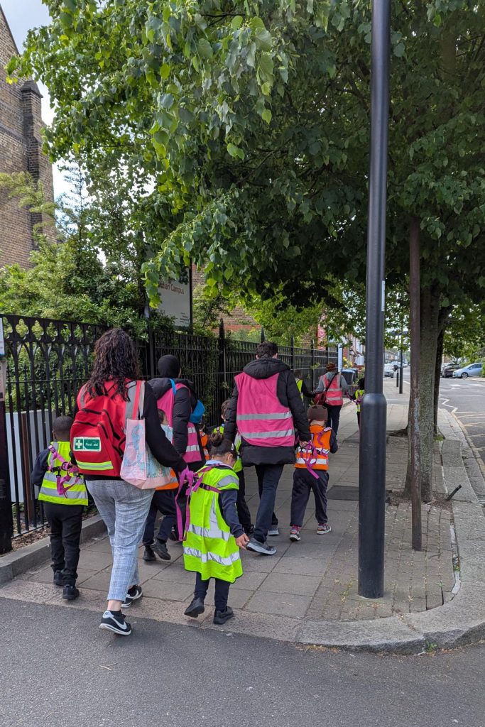 A group of adults and young children, some wearing bright safety vests, walk together on a pavement beside a metal fence and trees on a city street.