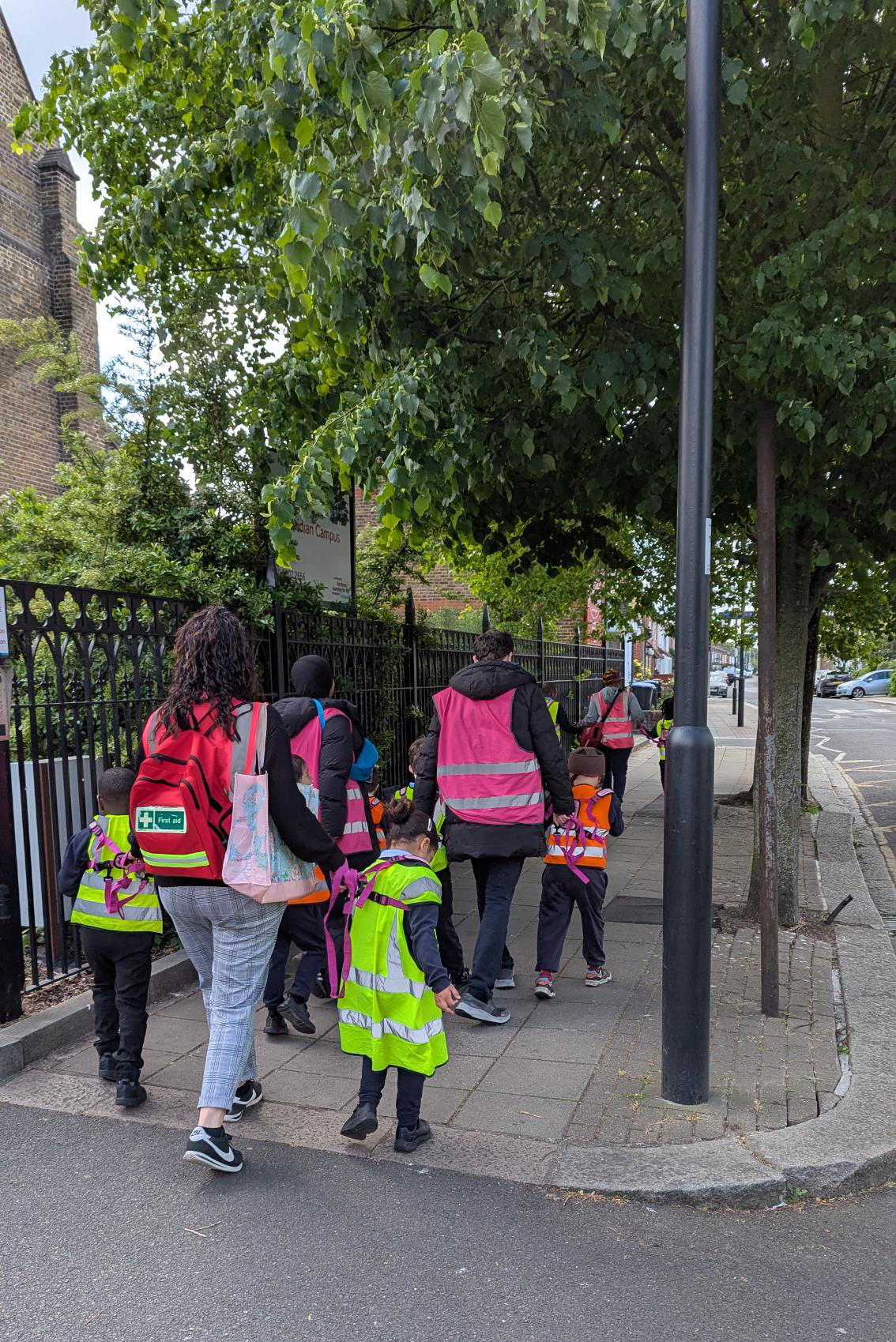 A group of adults and young children, some wearing bright safety vests, walk together on a pavement beside a metal fence and trees on a city street.