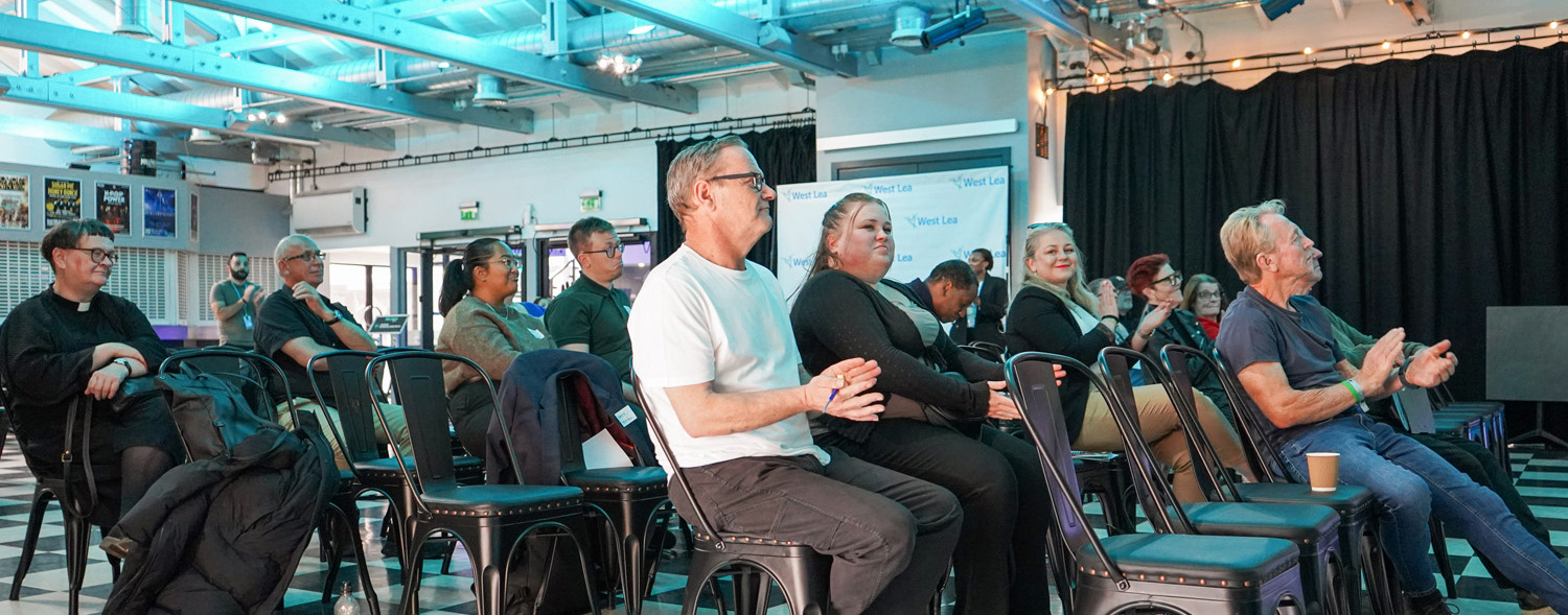 A group of people sit on chairs in a brightly lit room, some applauding and others listening attentively, with black curtains and a screen in the background. The atmosphere appears to be that of a casual event or presentation.