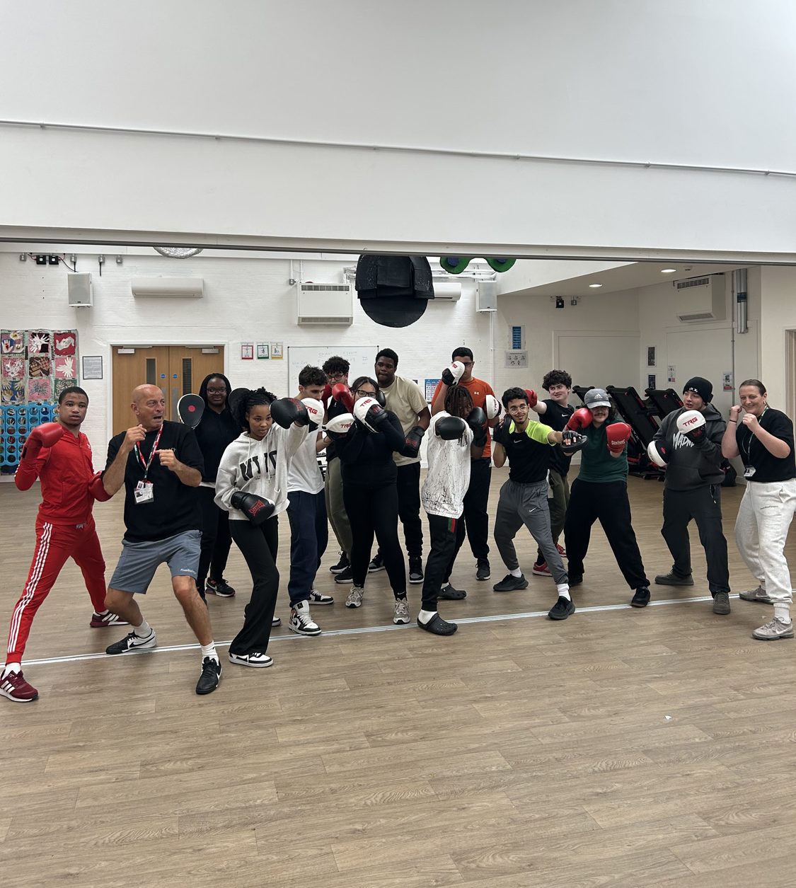 A group of people pose in a boxing stance inside a gymnasium with wooden floors and mirrors. Most are wearing sports clothing and boxing gloves, and they look ready for a boxing session.