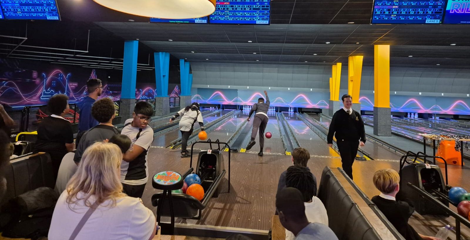 A group of people are at a bowling alley, watching and playing. Several lanes are in use, with colourful balls and bowling pins visible. Overhead screens display scores, and the area is brightly lit with neon décor.