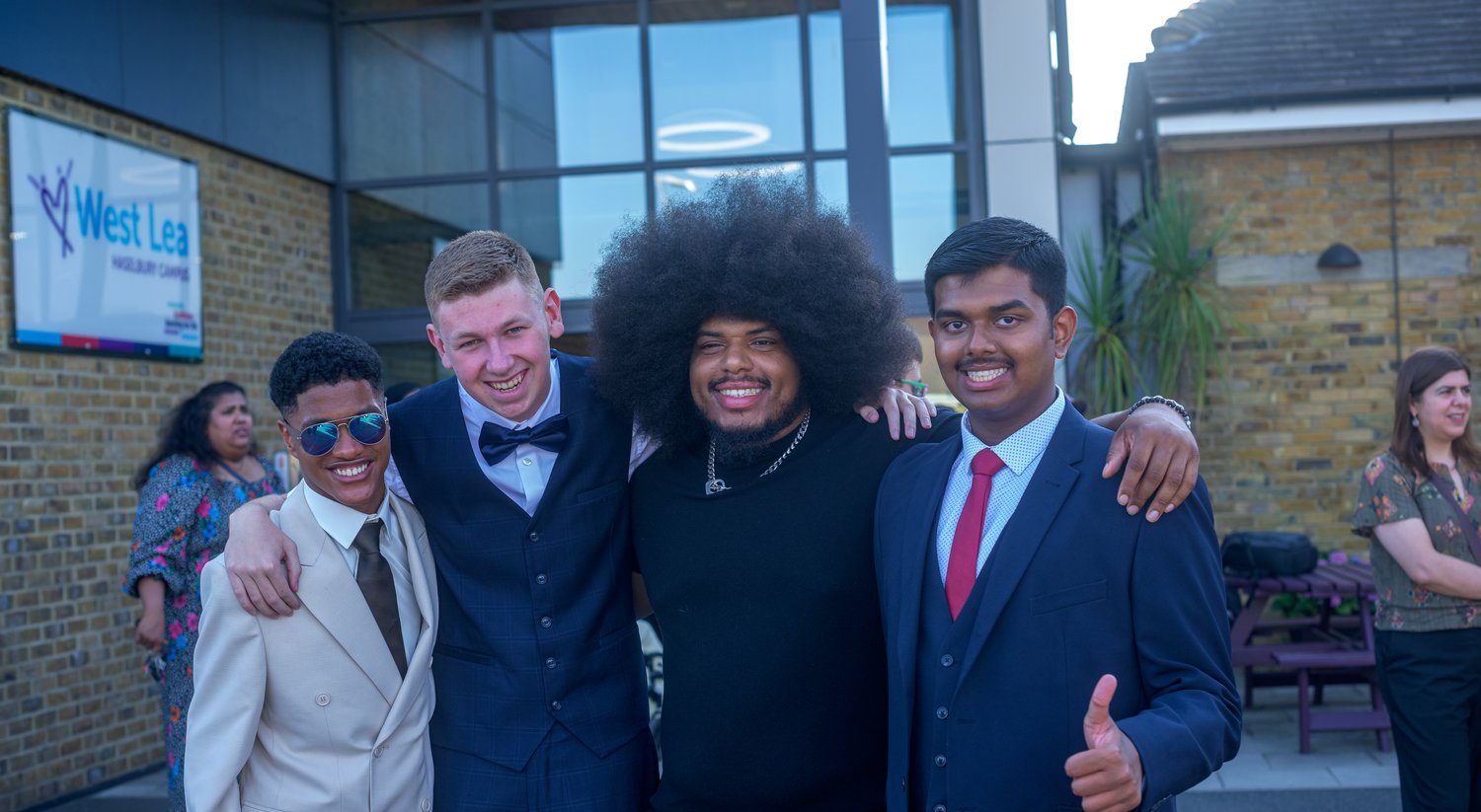 Four young men dressed in suits stand arm-in-arm, smiling outdoors in front of a building. One gives a thumbs up. Other people and a West Lea sign are visible in the background.