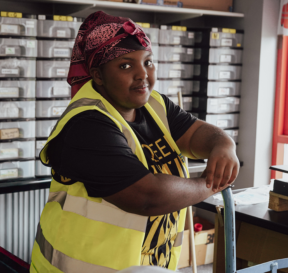 A person wearing a pink headscarf, yellow safety vest, and black T-shirt leans on a chair in a room with organised storage boxes and shelves in the background.