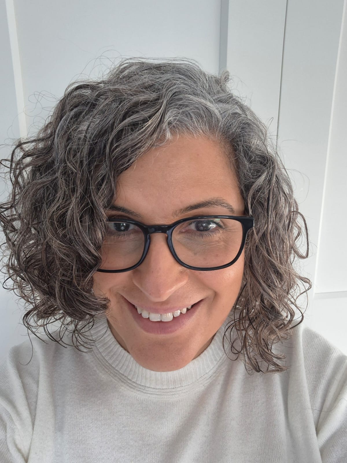 Smiling person with curly grey hair, wearing black glasses and a white jumper, standing in front of a white background.