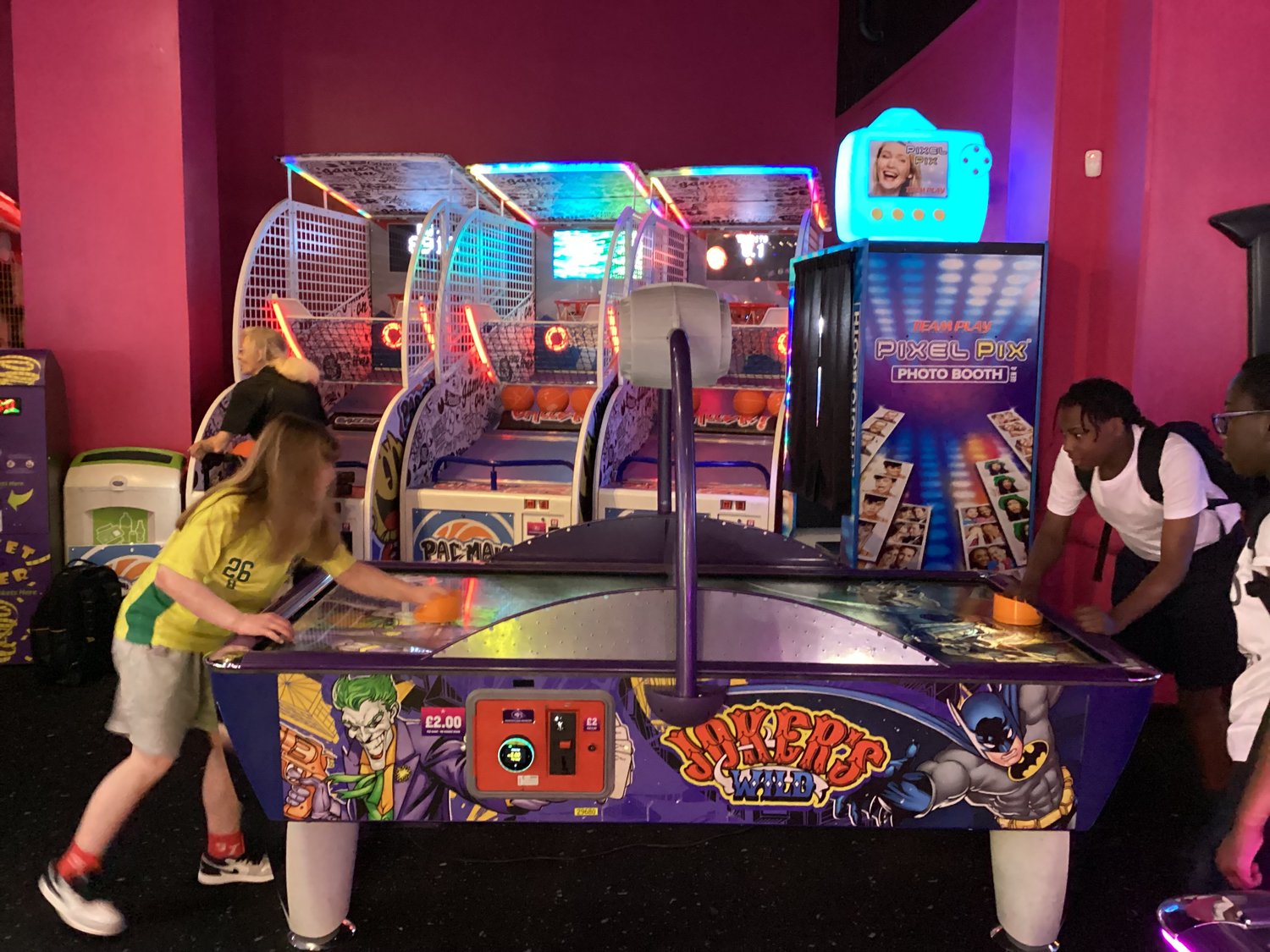 Three people play air hockey on a purple Joker-themed table in an arcade, with bright neon basketball games and a photo booth in the background. One player is striking the puck whilst the others watch closely.