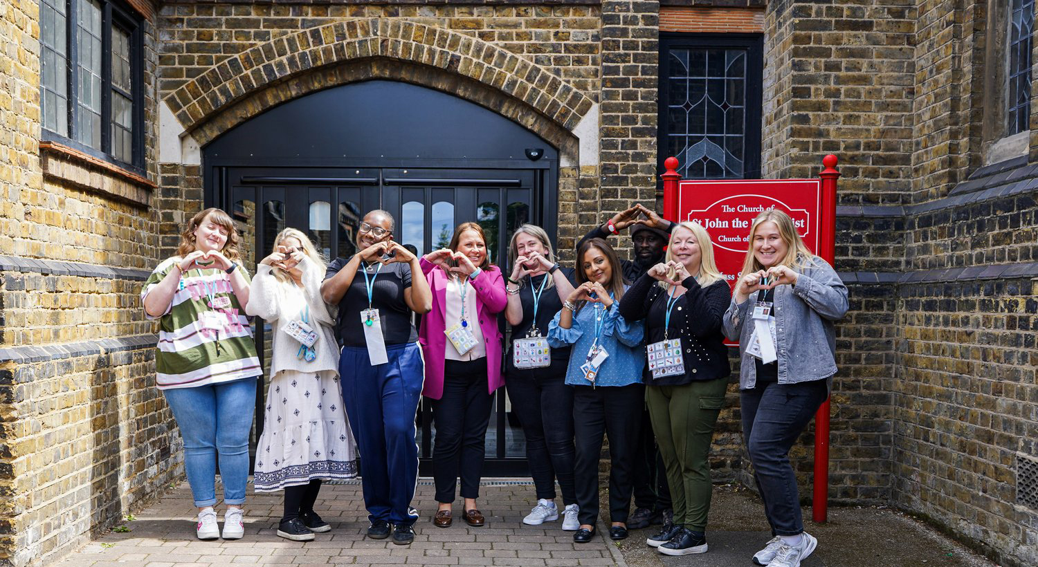 A group of ten people stand in front of a brick building, smiling and making heart shapes with their hands. They wear name badges and lanyards, and there is a red sign on the right side of the image.
