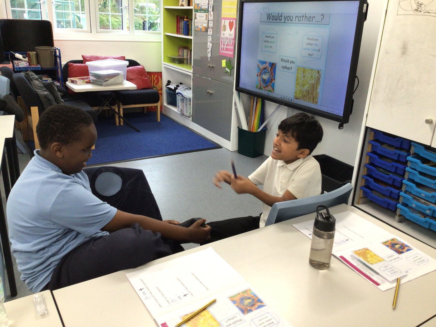 Two boys sit at a classroom table, smiling and talking. Behind them is a screen with Would you rather...? shown. Papers, pens, and a water bottle are on the table. Shelves and classroom materials are in the background.