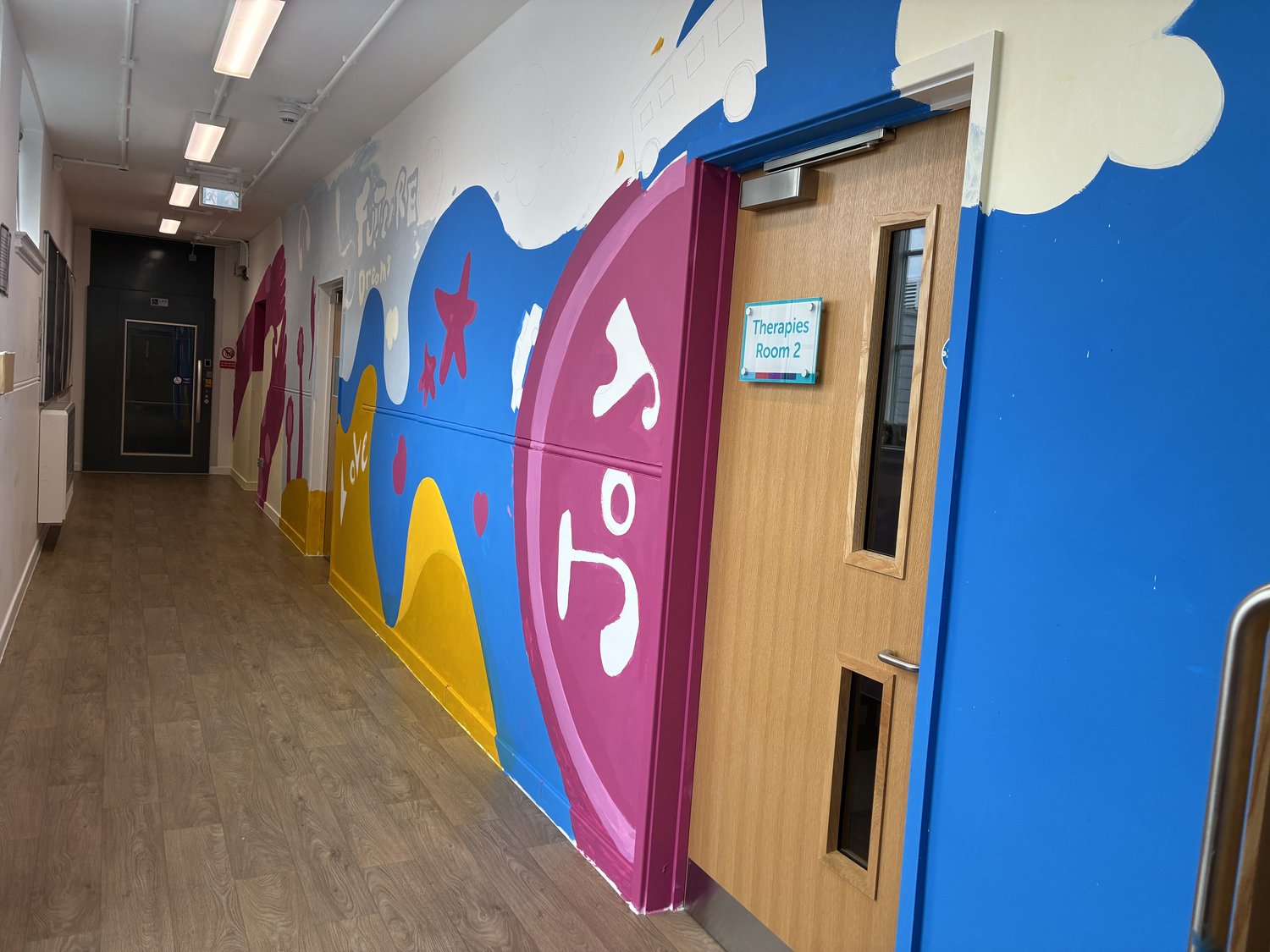 A school corridor with wood-look flooring and a colourful mural featuring blue, yellow, and magenta shapes, leading to a wooden door with a sign labelled Therapies Room 2.